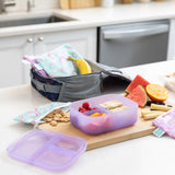 A kitchen counter with Bumkins Lunchbox Legends 6-Piece Set: Unicorns Purple Jelly lunch bag, cutting board, and fruit & veggies.