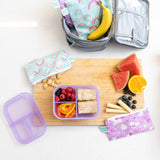 A kitchen counter with Bumkins Lunchbox Legends 6-Piece Set: Unicorns Purple Jelly lunch bag, cutting board, and fruit & veggies.