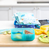 A kitchen counter with Bumkins Lunchbox Legends 6-Piece Set: Ocean Blue Jelly lunch bag, cutting board, and fruit & veggies.