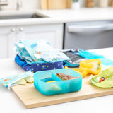 A kitchen counter with Bumkins Lunchbox Legends 6-Piece Set: Ocean Blue Jelly lunch bag, cutting board, and fruit & veggies.