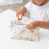 A child in a white shirt holds Bumkins Reusable Snack Bag, Small 2 Pack: Palm Check while reaching for a snack at a white table.