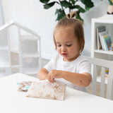 A toddler opens a Bumkins Reusable Snack Bag, Small 2 Pack: Palm Check at a white table with another bag nearby; a plant & shelves sit behind.