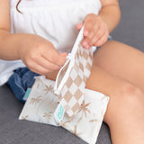 A child in a white top and blue shorts opens a Bumkins Reusable Snack Bag, Small 2 Pack: Palm Check on a gray couch.