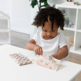 A curly-haired child at a table reaches inside a Bumkins Reusable Snack Bag, Small 2 Pack: Palm Check at a white table with another bag nearby.