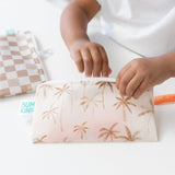 A child's hand reaches into a Bumkins Reusable Snack Bag, Small 2 Pack: Palm Check on a white table.