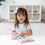 A smiling girl with a white bow holds a Bumkins Reusable Snack Bag, Small 2 Pack: Double Check, at a white table with a shelf behind her.