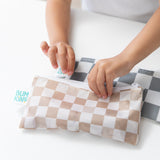 Childs hands opening a checkered zippered pouch labeled Bumkins. Another pouch with a similar pattern is nearby on a white surface.