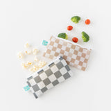 Bumkins Reusable Snack Bag, Small 2 Pack: Double Check amidst broccoli, cherry tomatoes, and popcorn on a white background.