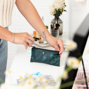A person zips a Bumkins Reusable Snack Bag, Small 2 Pack: Desert Night on a white table, surrounded by flowers and makeup.
