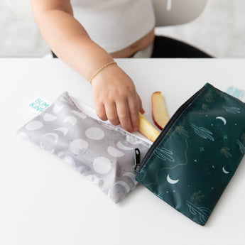 A child's hand reaches for apple slices from Bumkins Reusable Snack Bag, Small 2 Pack: Desert Night on a white table.