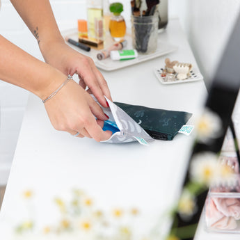A person packs toiletries into Bumkins Reusable Snack Bag, Small 2 Pack: Desert Night on a white countertop surrounded by flowers and makeup.