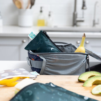 A Bumkins Reusable Snack Bag, Small 2 Pack: Desert Night sits in a lunchbox with a banana on a kitchen counter surrounded by food and bags.