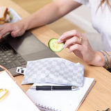 A person on a laptop holds a cucumber slice over a Roamio by Bumkins Reusable Snack Bag, Small 2 Pack: Rich & Boujee by a notepad & pen on a desk.