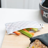A Roamio by Bumkins Reusable Snack Bag, Small 2 Pack: Rich & Boujee holds carrot & celery sticks on a cutting board on a kitchen counter.