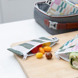 Small tomatoes spill from a Roamio by Bumkins Reusable Snack Bag, Small 2 Pack: Motley Club on a cutting board in front of a lunch bag.