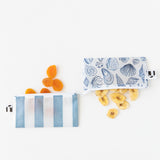 Dried apricots and banana slices spill from Roamio by Bumkins Reusable Snack Bag, Small 2 Pack: Coastal on a white backgorund.