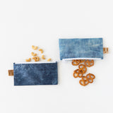 Cashews and pretzels spill out of Roamio by Bumkins Reusable Snack Bag, Small 2 Pack: Denim on Denim on a white background.