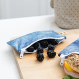 Blackberries spill from a Roamio by Bumkins Reusable Snack Bag, Small 2 Pack: Denim on Denim on a wood cutting board on a kitchen counter.