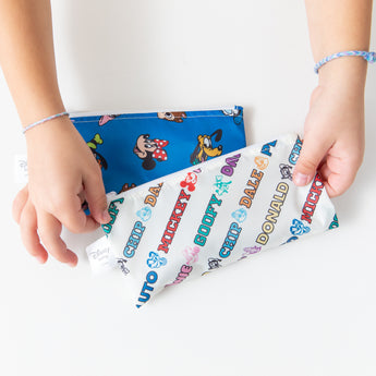 Hands hold Bumkins Reusable Snack Bag, Small 2 Pack: Mickey & Friends MVP at a white table.