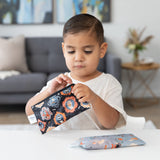 A young child clutches a Bumkins Reusable Snack Bag, Small 2 Pack: Everyday Is Halloween at a table, with a couch and plant in the background.