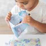 A toddler in a white shirt examines snacks in a Bumkins Reusable Snack Bag, Small 2 Pack: Stitch Star-ring Up Trouble at a white table.