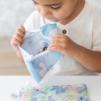A toddler in a white shirt examines snacks in a Bumkins Reusable Snack Bag, Small 2 Pack: Stitch Star-ring Up Trouble at a white table.
