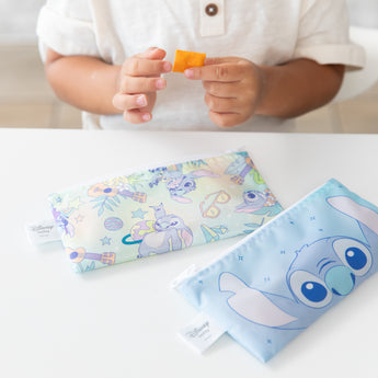 A child holds a cracker above Bumkins Reusable Snack Bag, Small 2 Pack: Stitch Star-ring Up Trouble on a white table.