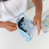 A child holds a Bumkins Reusable Snack Bag, Small 2 Pack: Stitch Star-ring Up Trouble with blueberries over a white table.
