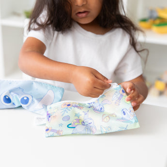 A child in white shirt unzips a Bumkins Reusable Snack Bag, Small 2 Pack: Stitch Star-ring Up Trouble at a white table.