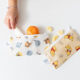Close-up of a child's hand grabbing snacks from Bumkins Reusable Snack Bag 4 Pack: Disney Pooh Bear + Friends on a white table.
