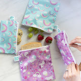 Bumkins Reusable Snack Bag 4 Pack: Unicorns and Rainbows lays on a marble surface with fruit and a sandwich; a hand unzips one of the bags.