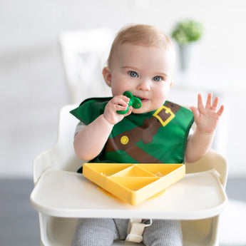 A baby in a high chair wears a Bumkins Caped SuperBib®: The Legend of Zelda™ Link, chewing a green spoon near a yellow triangle plate.