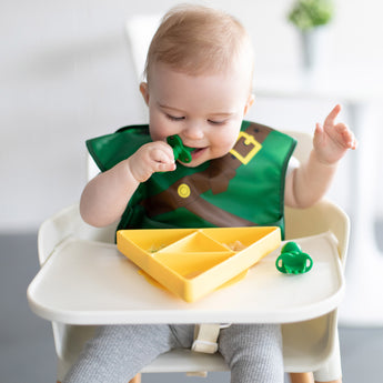 A baby in a Bumkins Caped SuperBib®: The Legend of Zelda™ Link sits in a high chair, eating from a yellow divided plate with green utensils.
