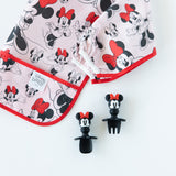 Bumkins Silicone Chewtensils®: Minnie Mouse rest next to a folded Minnie Mouse-patterned bib on a white background.