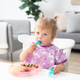 In a cozy living room, a toddler in a purple unicorn bib uses Bumkins Silicone Chewtensils®: Blue Jelly for baby-led weaning.