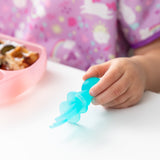 Close-up of a child holding Bumkins Silicone Chewtensils®: Blue Jelly near a pink sectioned plate, dressed in a unicorn-patterned bib.