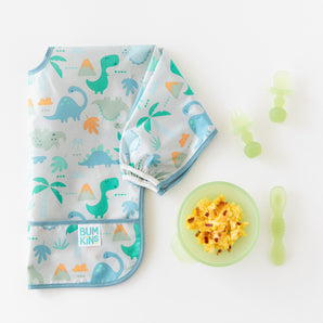 A folded dinosaur-print bib lays next to a green bowl with eggs and Bumkins Silicone Chewtensils®: Green Jelly on a white background.