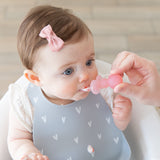 A baby in a gray Bumkins Silicone Bib 2 Pack: Hearts and Rainbows is fed by an adult hand with a pink spoon.