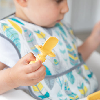 A young child in a colorful bib holds a Bumkins Silicone Chewtensils®: Pineapple.