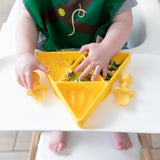 A baby eats from a yellow triangle plate with Bumkins Silicone Chewtensils®: Pineapple nearby on the high chair tray.