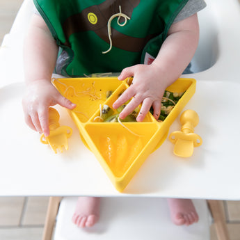 A baby eats from a yellow triangle plate with Bumkins Silicone Chewtensils®: Pineapple nearby on the high chair tray.