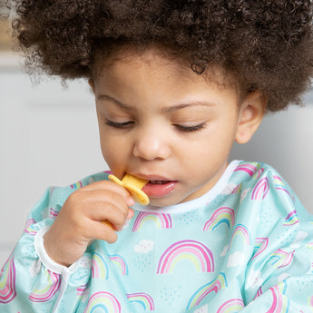 A toddler in a rainbow-patterned bib chews on a Bumkins Silicone Chewtensils®: Pineapple.