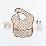 Bumkins Silicone Chewtensils®: Sand sit to the left of a tan checkered bib on a white background., with another set of utensils to the right.