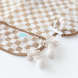 Close-up of Bumkins Silicone Chewtensils®: Sand on a white background next to a tan checkered bib.
