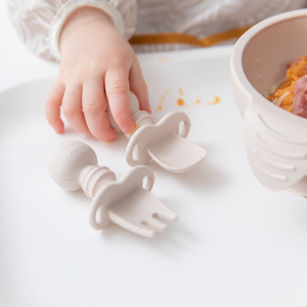 Close-up of a child's hand grabbing Bumkins Silicone Chewtensils®: Sand on a high chair tray with a bowl of food nearby.