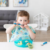 A young child uses Bumkins Spoon + Fork in Dark Blue and eats from a sectioned blue plate while wearing a whale bib.