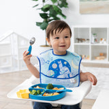 A toddler smiles in a high chair, eating with Bumkins Spoon + Fork: Dark Blue and a plate of food, wearing a blue space bib.