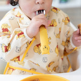 Close-up of a child in a yellow bear-patterned bib holding a Bumkins Spoon + Fork: Winnie the Pooh in their mouth.