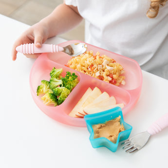 A child at a table, wearing a white shirt enjoys a meal from a Bumkins Silicone Grip Dish: Pink Jelly with a Little Dipper Aqua Jelly attached.