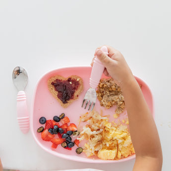A child's hand holds a fork over a Bumkins Silicone Grip Tray: Pink with heart-shaped toast, fruit, and hash browns on a white table.